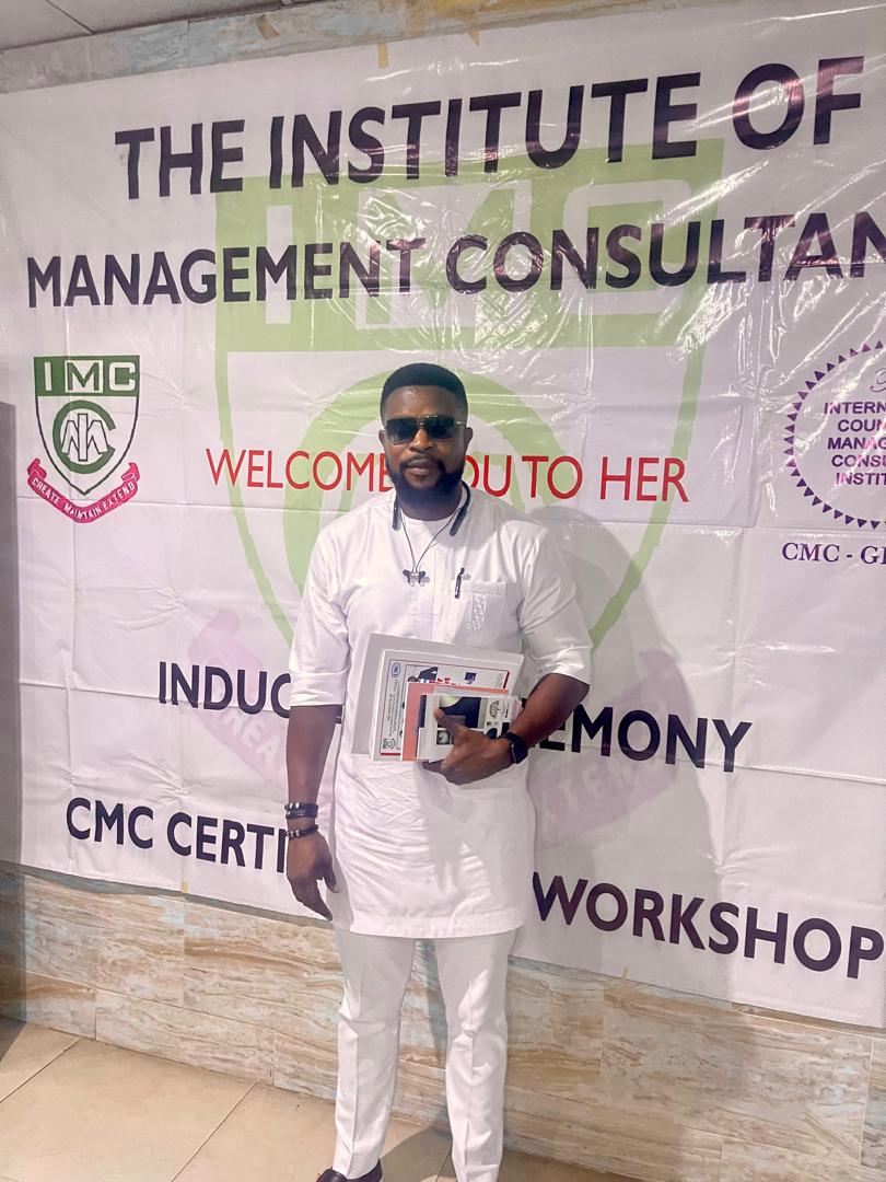 Victor in white traditional attire holding framed certificates before an Institute of Management Consultants banner